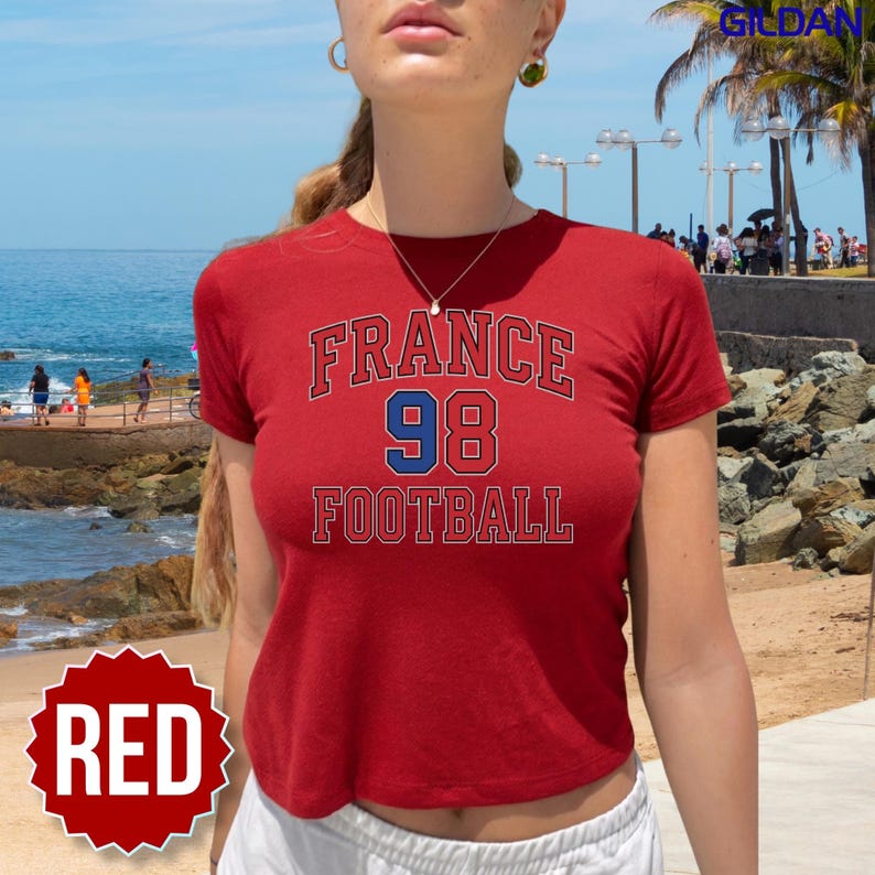 Close up of a woman wearing a vibrant red Gildan youth shirt styled as a 90s baby tee on a sunny beach boardwalk. Featuring the bold France 98 Football graphic, this top brings nostalgic Zidane era energy to your modern 2026 World Cup style. It is an ideal sportswear piece for hanging out with friends on match day, visiting local sports bars, or chanting in stadium tribunes, packaged with quick US domestic shipping for the ultimate summer look.