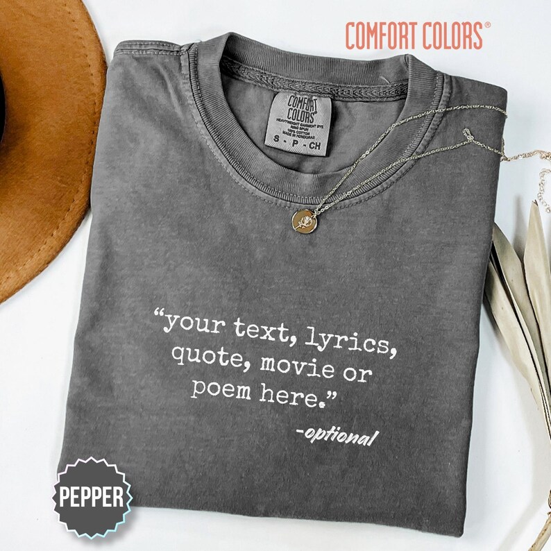以下が含まれることがあります： 「your text, lyrics, quote, movie or poem here.」およびその下に「-optional」と白でプリントされたチャコールグレーのComfort Colors製Tシャツ。小さな円形ペンダントが付いた繊細なゴールドトーンのネックレスがネックラインに置かれています。