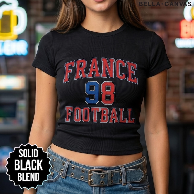 Midsection view of a black ribbed baby tee displaying the vintage inspired France 98 Football block letters. Styled with a double grommet belt and jeans in a sports bar atmosphere, this top combines 90s streetwear with 2026 soccer tournament spirit. It is designed for passionate fans to wear while shouting in the stands or enjoying drinks with a group, offering a subtle nod to the legendary 1998 squad performance with fast domestic US shipping.