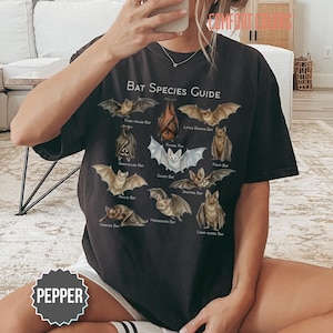 May include: A black t-shirt featuring a graphic titled Bat Species Guide with illustrations of various bat types including Free-tailed, Flying Fox, Little Brown, Spectacled, Ghost, Fruit, Pallid, Spotted, Vampire, Horseshoe, and Long-eared bats.