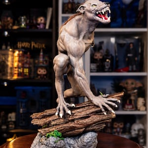 Könnte beinhalten: Eine detaillierte Werwolf-Figur in kauernder Haltung mit grauer Haut, scharfen weißen Krallen und einem geöffneten Maul mit sichtbaren Zähnen. Die Kreatur steht auf einem strukturierten braunen Holzstamm mit kleinen grünen Moosflecken und grauem Fels.