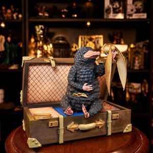 Puede incluir: Una figura de Niffler negro sentada dentro de una maleta marrón abierta. La criatura tiene un hocico largo, pelaje texturizado y pies rosados, sosteniendo una bola alada dorada en una mano mientras hay monedas de oro en su bolsa y maleta.