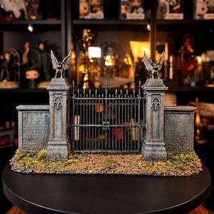 Puede incluir: Un modelo en miniatura de una puerta de cementerio con dos pilares color piedra rematados con estatuas de gárgolas aladas, unidos por una valla de metal negro. La base tiene textura de suelo marrón y pequeñas manchas de musgo verde.