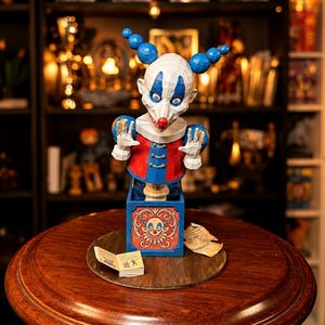 Può includere: Un jack-in-the-box in carta raffigurante un clown con viso bianco, segni blu e capelli a sfere blu. Il clown indossa una giacca rossa e blu con bottoni dorati. La base blu mostra una colorata illustrazione di un clown e piccoli libri di carta.