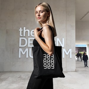 Pode incluir: Bolsa tote preta com texto branco "303 808 909". A bolsa é carregada por uma pessoa em frente a um edifício com as palavras "the DESIGN MUSEUM" em letras brancas grandes.
