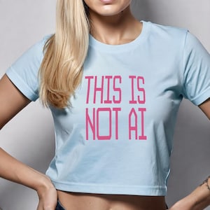 Op de afbeelding: Lichtblauw crop top met de roze tekst "THIS IS NOT AI". Het shirt heeft korte mouwen en een ronde hals. De stijl is casual en modern, geschikt voor dagelijks gebruik.