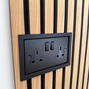 May include: A black double electrical socket and light switch set into a wooden panel. The panel features vertical wooden slats with black detailing. The wood has a light brown finish. The sockets are British standard.
