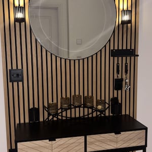 May include: A contemporary hallway arrangement with a circular mirror, wall lights, and a black console table with two drawers. The wall has a wooden slat design. Includes candle holders and key hooks.