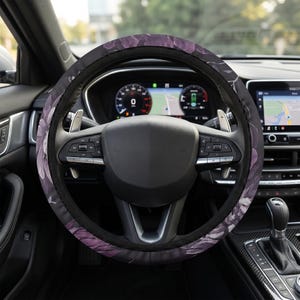 Puede incluir: Una funda para volante con un estampado floral en color morado oscuro y negro. La funda está instalada en un volante negro con levas de cambio plateadas y botones de control integrados, dentro del interior de un vehículo.