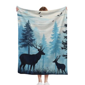 May include: A rectangular throw blanket featuring a printed forest scene with two dark silhouettes of deer standing in tall grass. The background displays light blue pine trees and a pale sky with several small black birds flying overhead.