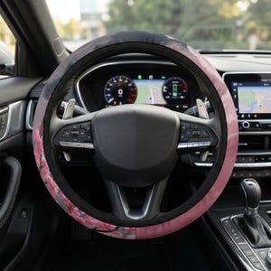 May include: A circular steering wheel cover featuring a printed design of pink cherry blossom branches against a dark grey and black background. The cover has a black fabric border along the inner and outer edges, fitted onto a car steering wheel.