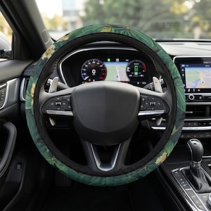May include: A steering wheel cover featuring a dark green background with a repeating pattern of large, light green tropical leaves and small, textured golden-yellow floral clusters, fitted onto a black car steering wheel with silver accents.