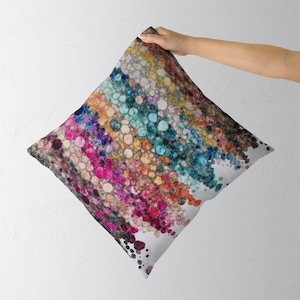 May include: A square throw pillow featuring a vibrant, abstract pattern of multicolored circular shapes. The design displays a gradient of pink, purple, orange, yellow, teal, and blue dots against a white background, creating a textured effect.