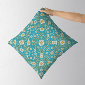 Op de afbeelding: Een vierkant sierkussen met een levendige turquoise blauwe achtergrond en een herhalend patroon van witte madeliefjes met gele harten, verweven met ingewikkelde, krullende goudgele filigraanversieringen over het gehele oppervlak.