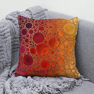May include: A square throw pillow featuring a vibrant gradient pattern of circular shapes. The design transitions from deep magenta and dark red on the left side to bright orange and warm yellow on the right, resembling a collection of bubbles.