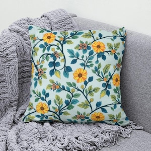 May include: A square throw pillow featuring a light blue background with a repeating pattern of bright yellow sunflowers, dark teal stems, and green leaves. The floral design covers the entire surface of the pillow, creating a dense botanical arrangement.