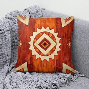 May include: A square throw pillow featuring a vibrant orange and deep red background with a central sun-like motif. The design includes a large cream-colored serrated circle surrounding concentric diamond shapes in shades of orange, red, and dark brown.