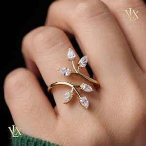 Könnte beinhalten: Ein goldfarbener offener Ring mit zwei verzweigten Stielen, die sich um den Finger legen. Jeder Stiel ist mit zwei klaren, birnenförmigen facettierten Steinen besetzt, die an Blätter erinnern. Das Design ist symmetrisch mit insgesamt vier Steinen.