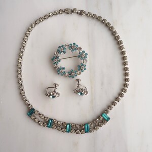May include: A vintage jewelry set with a delicate blue rhinestone necklace, matching earrings, and a brooch. The necklace features a double row of small rhinestones with larger rectangular stones interspersed throughout. The earrings and brooch are both circular and feature a cluster of smaller rhinestones.