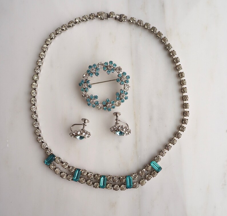 May include: A vintage jewelry set with a delicate blue rhinestone necklace, matching earrings, and a brooch. The necklace features a double row of small rhinestones with larger rectangular stones interspersed throughout. The earrings and brooch are both circular and feature a cluster of smaller rhinestones.