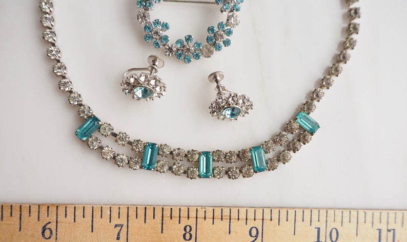 May include: A vintage jewelry set with a necklace, earrings, and a brooch. The set features clear and light blue rhinestones in a silver tone metal.