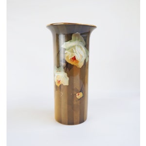May include: A tall, brown vase with vertical stripes and a gold rim. The vase is adorned with hand-painted white and yellow roses with red centers. The floral design adds a touch of elegance to this decorative piece.