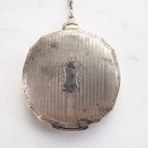 May include: A silver compact with a decorative design and a hinged lid. The compact has a chain attached to the top.