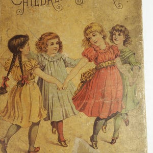 May include: Four young girls in dresses are holding hands and dancing in a circle. The girls are wearing yellow, blue, red, and green dresses. The background is a light brown color.