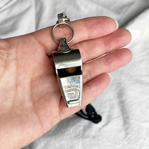 May include: Silver whistle with a black lanyard. The whistle is engraved with "The Acme Thunderer Made in England."