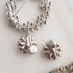 May include: A pair of silver tone clip-on earrings with a floral design. The earrings are marked "BOOCHE" on the back.