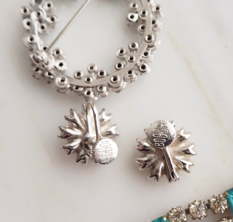 May include: A pair of silver tone clip-on earrings with a floral design. The earrings are marked "BOOCHE" on the back.