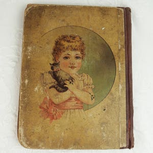 May include: A vintage children's book cover with a worn, yellowed paper cover. The cover features a young girl with blonde hair holding a black cat. The girl is wearing a white dress with a pink sash and a pink bow. The image is set against a green background.