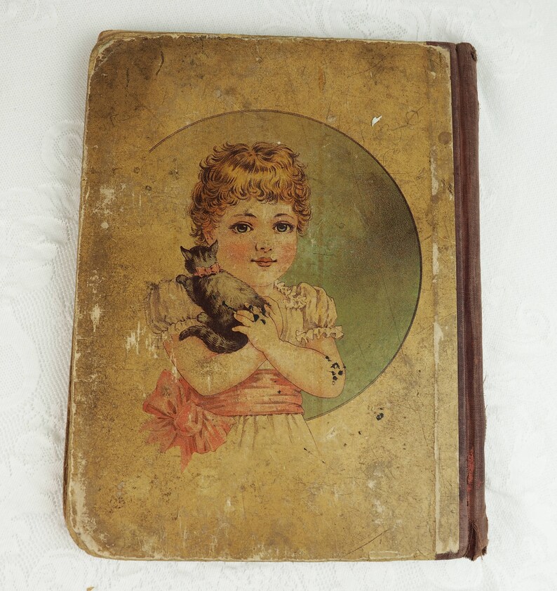 May include: A vintage children's book cover with a worn, yellowed paper cover. The cover features a young girl with blonde hair holding a black cat. The girl is wearing a white dress with a pink sash and a pink bow. The image is set against a green background.