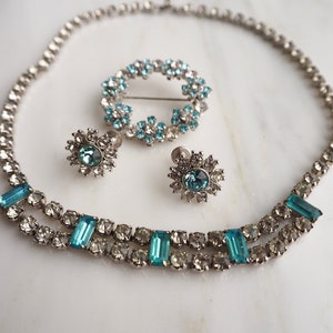 May include: A silver necklace with a double row of clear and blue rhinestones. The necklace is paired with matching earrings and a brooch, all featuring a similar design.