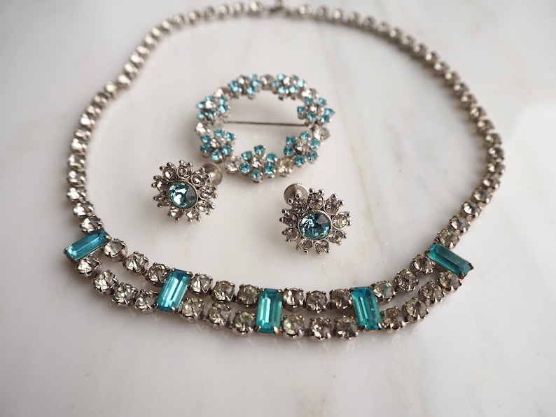 May include: A silver necklace with a double row of clear and blue rhinestones. The necklace is paired with matching earrings and a brooch, all featuring a similar design.