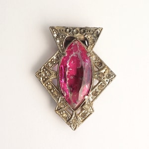 May include: A silver-toned brooch with a large, pink, faceted glass stone in the center. The stone is surrounded by a decorative, geometric design.