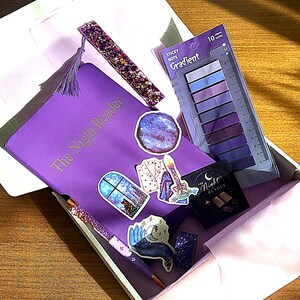 Peut inclure: Une boîte cadeau violette remplie d'articles de papeterie. Comprend un journal avec le texte "The Night Reader", des autocollants, un stylo, un marque-page et des notes adhésives dans les tons violets et bleus. Les articles sont disposés sur une surface en bois.