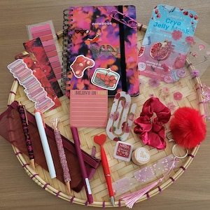 Peut inclure: Une collection d'articles de papeterie comprenant un carnet à spirales au motif rose et violet, un sachet de masque Cryo Jelly, un chouchou en satin rouge, un porte-clés pompon rouge, divers stylos, un bloc-notes rose marqué I BELIEVE IN YOU et des autocollants.