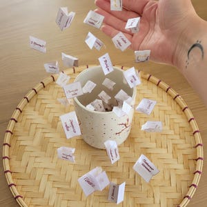 Peut inclure: Une tasse en céramique beige avec un motif floral est posée sur un plateau en bambou tressé. De petits papiers blancs avec du texte manuscrit sont éparpillés autour de la tasse. Le plateau a une bordure rouge foncé.
