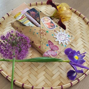 Peut inclure: Un sac cadeau décoratif avec des autocollants en forme d'étoiles et de papillons, posé sur un plateau tressé. Le sac contient un tube de Lipton Matcha, un stylo et d'autres articles. L'arrangement est orné de fleurs violettes et d'un iris jaune et violet.