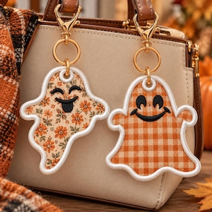 May include: Two ghost-shaped bag charms with gold-tone metal lobster clasps and rings. One ghost features an orange floral pattern with a black embroidered face, while the other displays an orange and white gingham check with a black smiling face.
