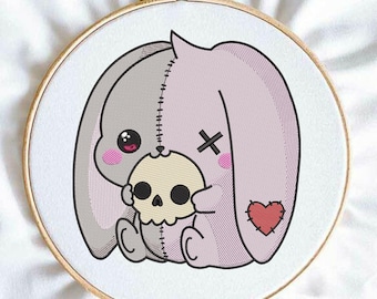 May include: An embroidery hoop containing a stitched design of a split-colored rabbit, half grey and half light purple, holding a small beige skull. The rabbit features a black X over one eye, pink cheeks, and a red heart patch on its side.