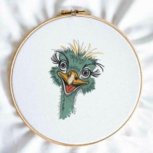 May include: Embroidered hoop art depicting a cartoon ostrich head. The ostrich has blue eyes, long eyelashes, and a yellow beak. The embroidery uses green, yellow, and red threads, set against a white fabric background.