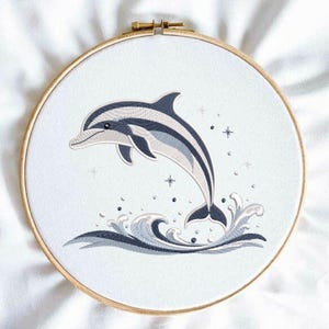 以下が含まれることがあります： 木製のフレームに刺繍されたイルカのデザイン。イルカは水から飛び出し、青と白のストライプ模様です。波と細部は青とグレーの色合いです。