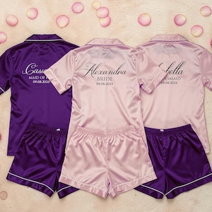May include: Three sets of satin pajama sets in purple and pink. Each set includes a short-sleeved button-down shirt and matching shorts. The shirts are personalized with names and dates. White trim accents the edges of the shirts and shorts. Scattered rose petals complete the scene.