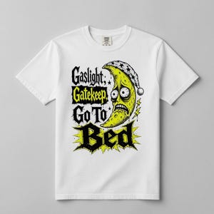 Peut inclure: T-shirt blanc avec un motif vieilli. Il présente une lune jaune portant un bonnet de nuit, avec les mots "Gaslight, Gatekeep, Go To Bed" en noir et jaune. La lune a une expression inquiète.