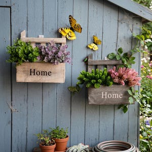 Puede incluir: Dos jardineras de madera con la palabra "Home" impresa, montadas en una pared azul desgastada, llenas de vegetación y flores moradas y rosas. Decoraciones de mariposas amarillas realzan el tema del jardín.