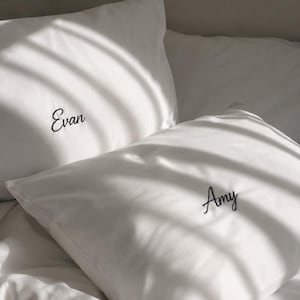 Op de afbeelding: Twee witte kussenslopen met aangepast borduurwerk in een zwart cursief lettertype. Op de ene kussensloop staat de naam Evan, terwijl op de andere kussensloop de naam Amy staat. De stof is glad en effen, met zachte schaduwen op het oppervlak.