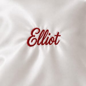 Op de afbeelding: Een wit satijnen stoffen oppervlak met de naam Elliot geborduurd in een rood cursief lettertype in het midden. Het materiaal heeft een gladde, reflecterende glans met zachte plooien die vanuit de geborduurde tekst naar buiten stralen.
