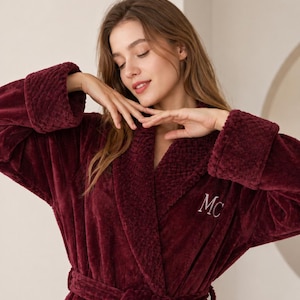 May include: A plush, burgundy-colored robe with a shawl collar and a matching belt. The robe features a textured design on the cuffs and collar, and the initials "MC" are embroidered on the chest in white.